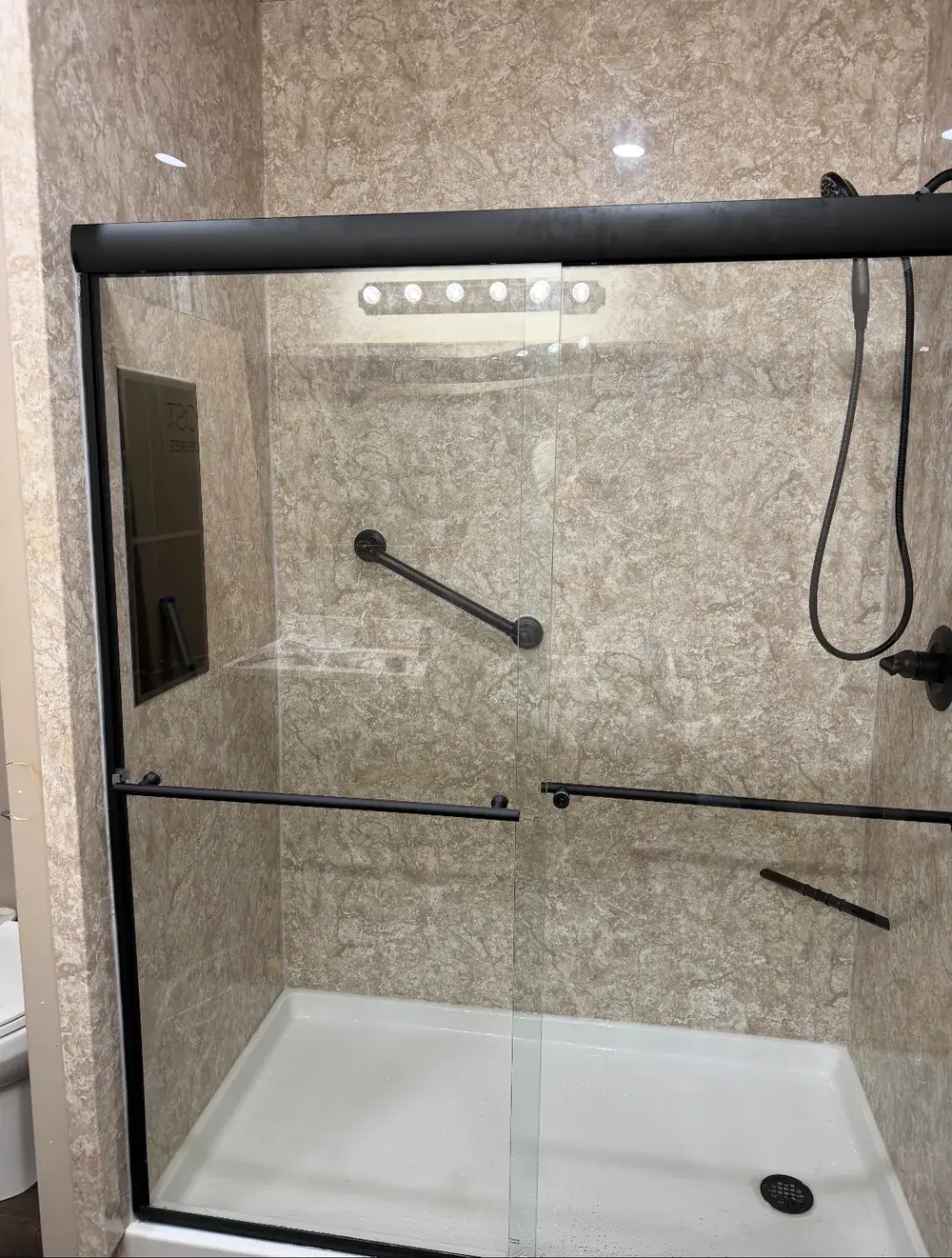 A sliding glass shower door with matte-black hardware over light stone-look wall panels and a built-in grab bar