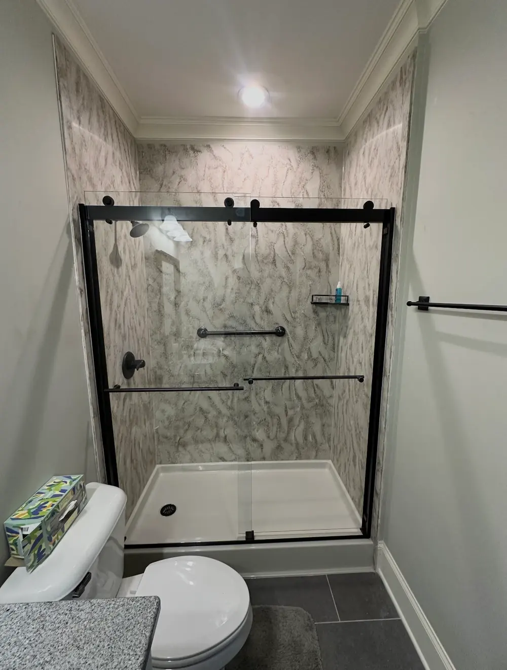 A frameless sliding shower door with matte-black hardware, stone-look wall panels, and an adjustable shelf caddy